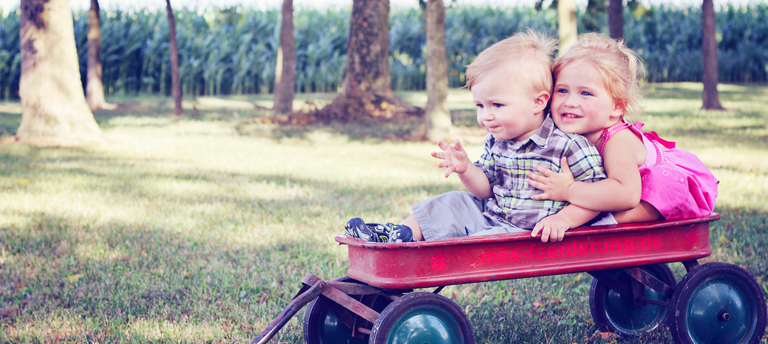 Zwei kleine Kinder sitzen auf einem roten Bollerwagen, der mit mbsfoerderung.de beschriftet ist. Das Bild symbolisiert das gesellschaftliche Engagement der Sparkasse.